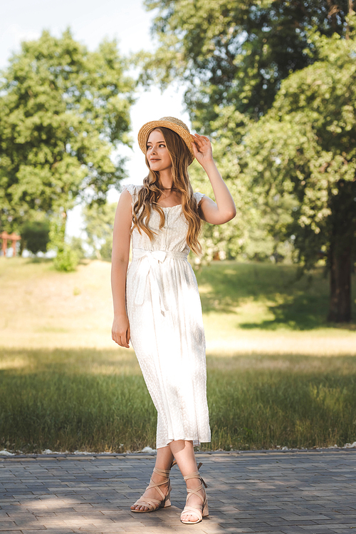 full length view of beautiful girl in white dress and straw hat standing on meadow and looking away