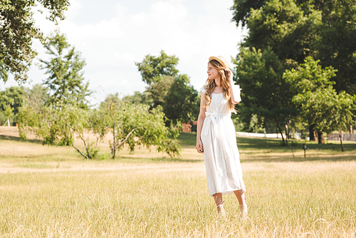 beautiful girl in white dress and straw hat standing on meadow and looking away