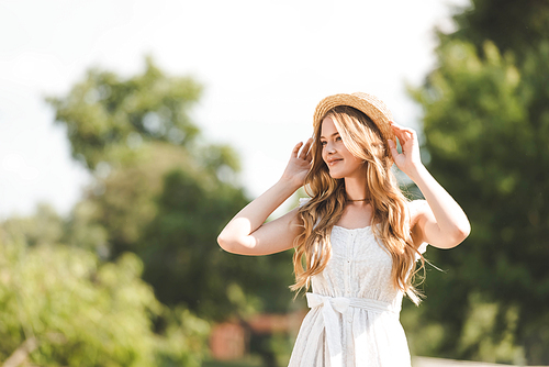 beautiful girl in white dress touching straw hat and smiling while standing on meadow and looking away