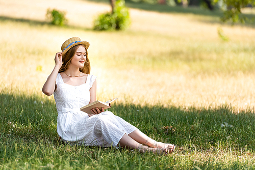 beautiful girl in white dress touching straw hat and holding book while sitting on meadow with closed eyes