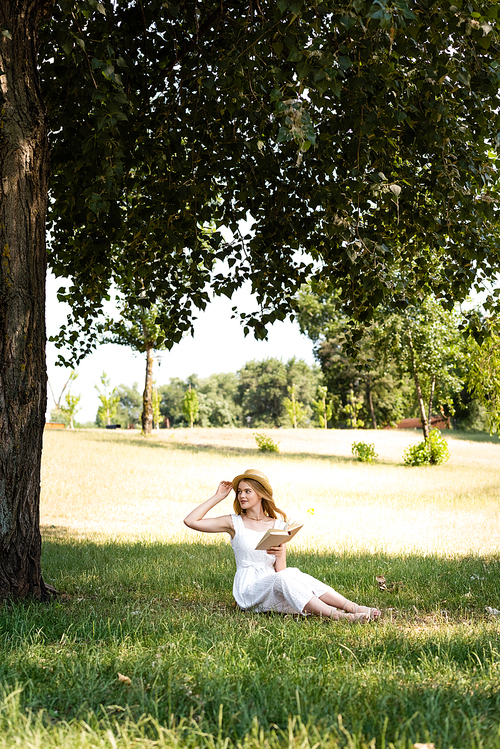 full length view of beautiful girl in white dress touching straw hat and holding book while sitting on meadow and looking away