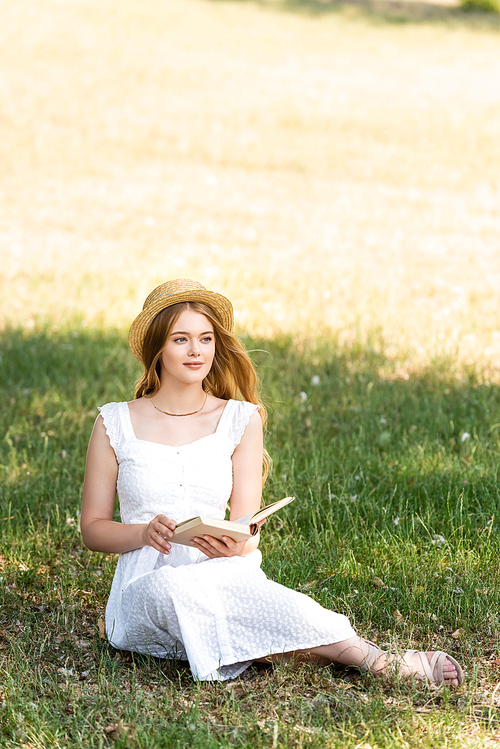 full length view of beautiful girl in white dress and straw hat holding book while sitting on meadow and looking away