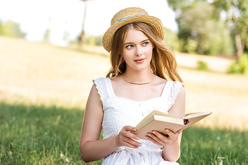 beautiful girl in white dress and straw hat holding book while sitting on meadow and looking away