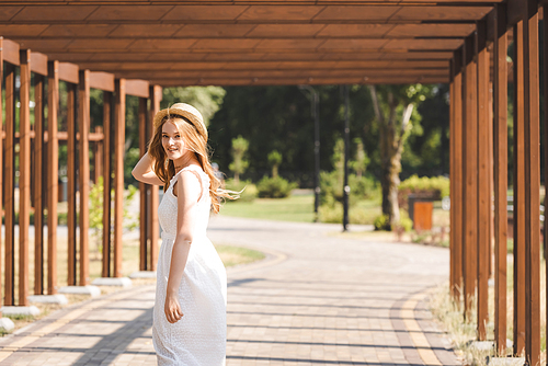 beautiful girl in white dress touching straw hat while walking near wooden construction and 