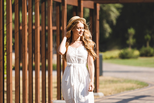 beautiful girl in white dress touching straw hat and smiling with closed eyes