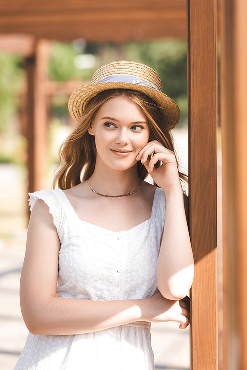 beautiful girl in white dress and straw hat leaning on wooden construction