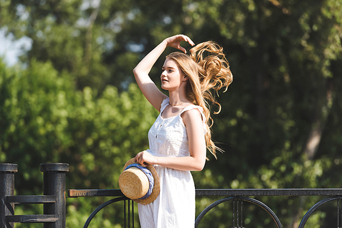 beautiful girl in white dress holding straw hat while touching hair and looking away