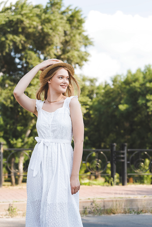 beautiful young girl in white dress touching straw hat while smiling and looking away