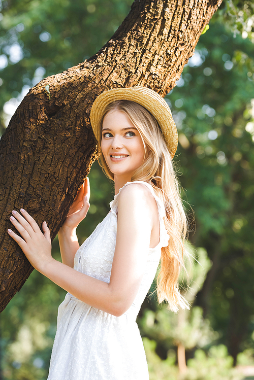 beautiful girl in white dress and straw hat standing near trunk of tree