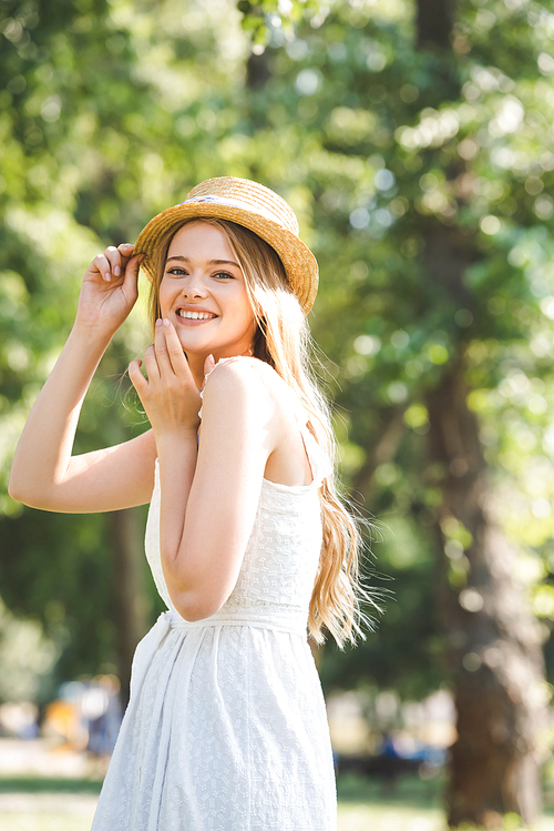 selective focus of beautiful young woman in white dress and straw hat smiling and 
