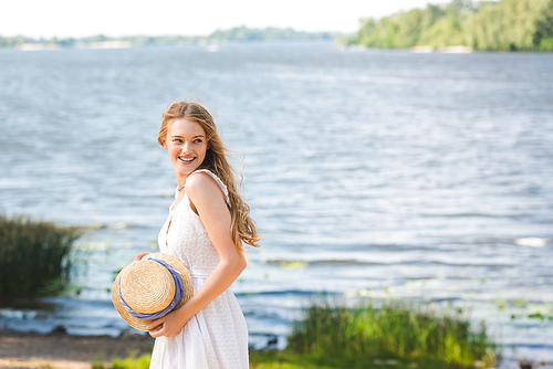 young girl standing on shore of river while holding straw hat and looking away