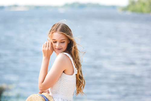 young girl standing on shore of river while holding straw hat and looking down