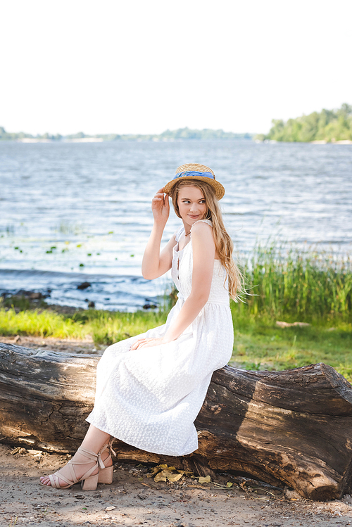 beautiful girl in white dress and straw hat sitting on trunk of tree on river shore and looking away