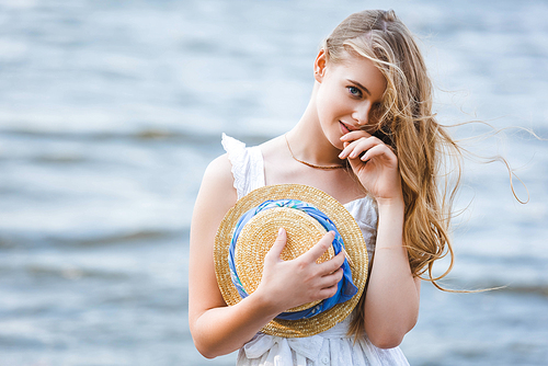 beautiful girl standing on river shore, holding straw hat, smiling and 