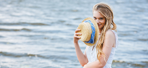 panoramic shot of beautiful girl holding straw hat near face, smiling and 