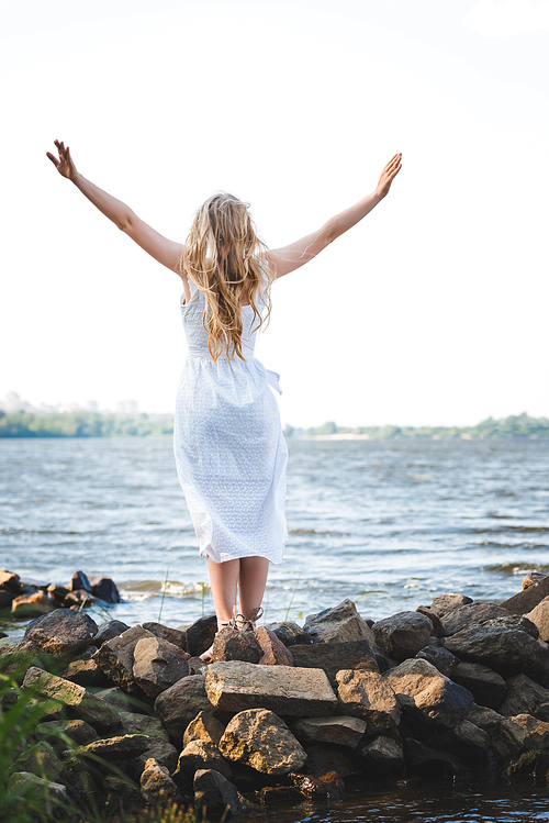 back view of girl in white dress standing on rocky river shore with hands in air