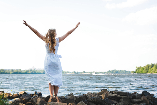 full length view of girl in white dress standing on rocky river shore with hands in air