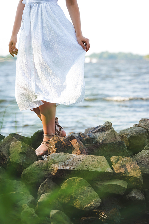 cropped view of girl in white dress walking on rocky river shore