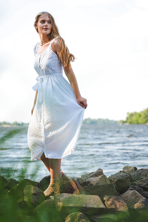 selective focus of beautiful girl in white dress walking on rocky river shore and looking away