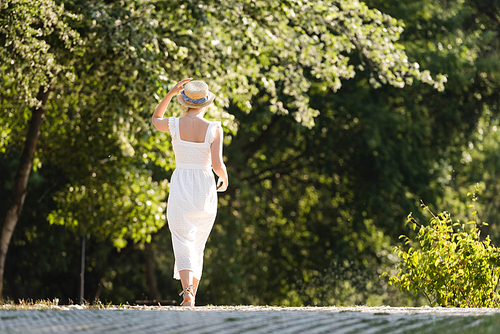 back view of girl in white dress touching straw hat while walking in park