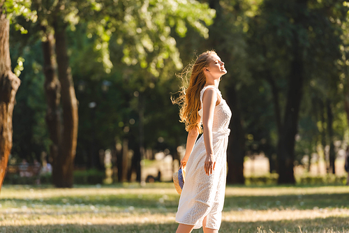 side view of beautiful girl in white dress holding straw hat while smiling and standing on meadow with closed eyes