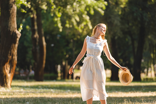 beautiful girl in white dress holding straw hat while smiling and standing on meadow with closed eyes