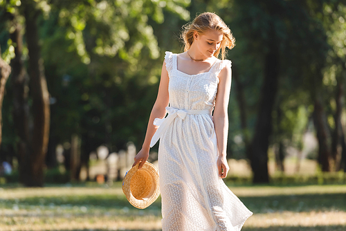 beautiful girl in white dress holding straw hat while smiling and standing on meadow and looking down
