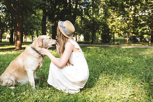 girl in white dress and straw hat petting golden retriever while sitting on meadow