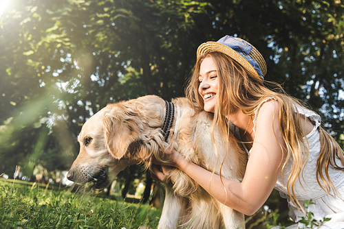 beautiful young girl in white dress and straw hat hugging golden retriever while smiling, sitting on meadow and looking at dog