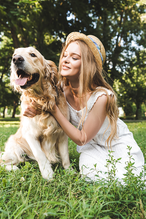 beautiful girl in white dress and straw hat petting golden retriever while smiling, sitting on meadow and looking at dog