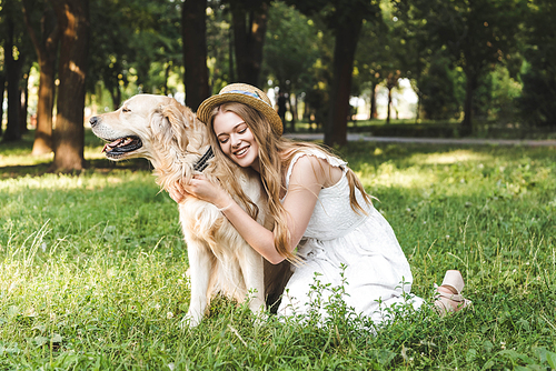 beautiful young girl in white dress and straw hat hugging golden retriever while smiling and sitting on meadow with closed eyes