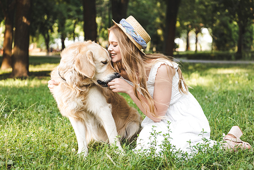beautiful young girl in white dress and straw hat feeding golden retriever while smiling and sitting on meadow with closed eyes