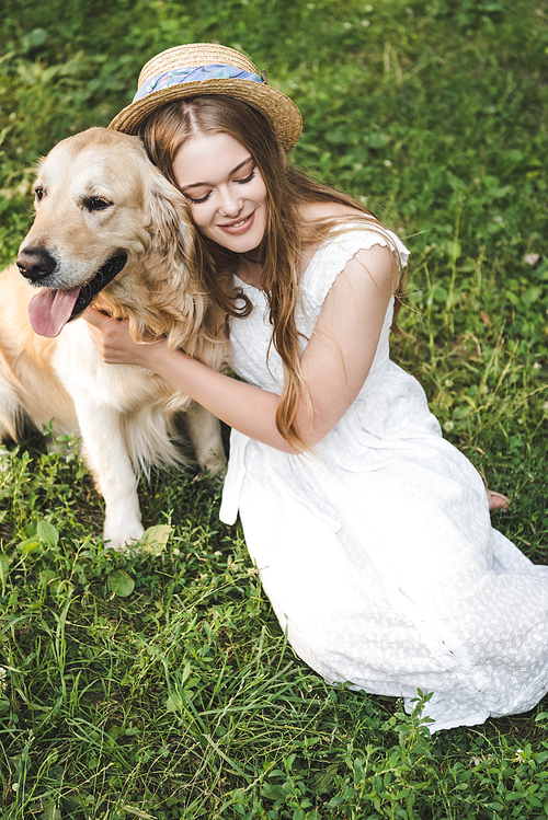high angle view of beautiful young girl in white dress and straw hat hugging golden retriever while smiling and sitting on meadow with closed eyes
