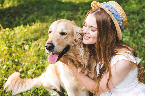 beautiful girl in white dress and straw hat hugging golden retriever while sitting on meadow and looking at dog