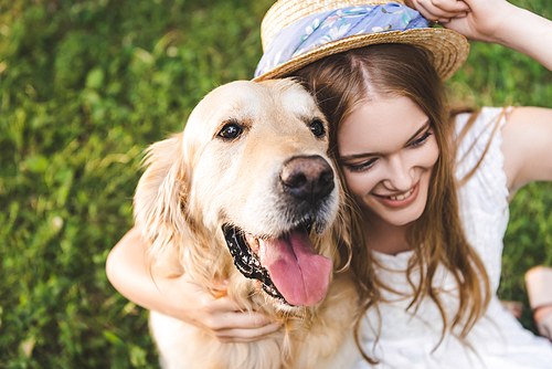 high angle view of beautiful young girl in white dress and straw hat hugging golden retriever while sitting on meadow, smiling and looking away