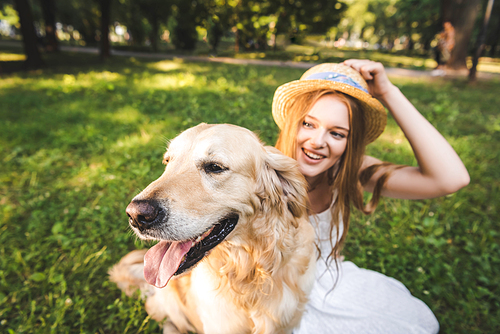 selective focus of beautiful young girl in white dress and straw hat hugging golden retriever while smiling, sitting on meadow and looking at dog