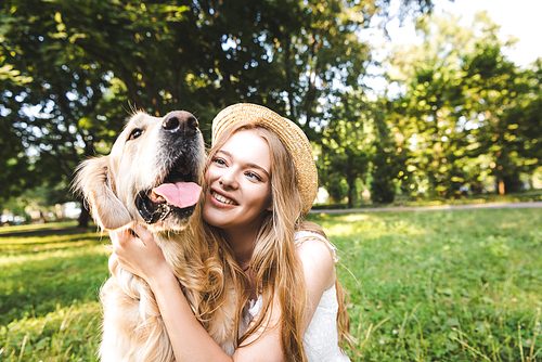 beautiful girl in white dress and straw hat hugging golden retriever while sitting on meadow, smiling and looking at dog