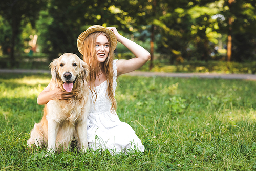 beautiful young girl in white dress touching straw hat while hugging golden retriever while smiling, sitting on meadow and looking away