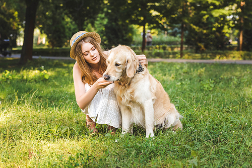 full length view of beautiful girl in white dress and straw hat petting golden retriever while sitting on meadow and looking at dog