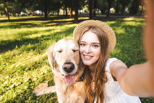 young girl in white dress and straw hat hugging golden retriever and taking selfie while sitting on meadow, smiling and 