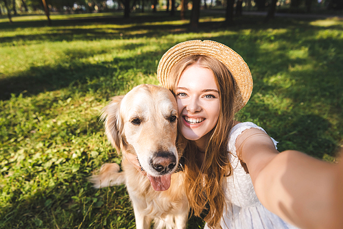 beautiful young girl in white dress and straw hat hugging golden retriever and taking selfie while sitting on meadow, smiling and 