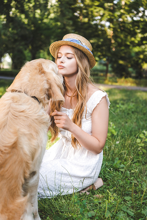 beautiful girl in white dress and straw hat petting golden retriever while sitting on meadow and looking at dog