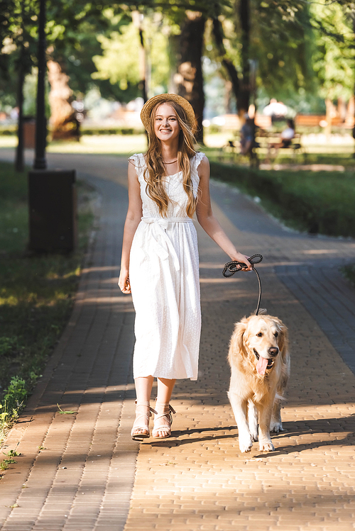 full length view of beautiful girl in white dress and straw hat walking with golden retriever on pathway and 