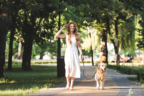 full length view of beautiful girl in white dress and straw hat walking with golden retriever on pathway and looking away