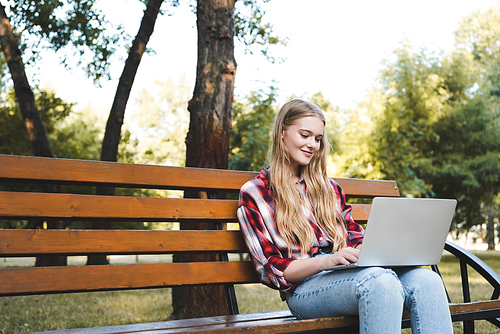 beautiful girl in casual clothes sitting on wooden bench in park and using laptop