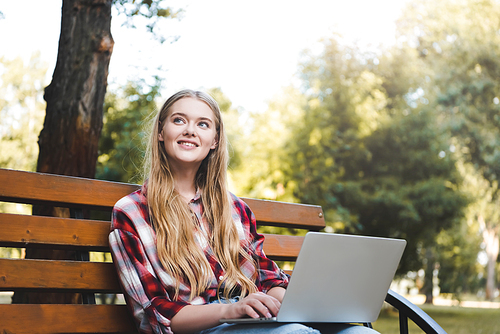 beautiful girl in casual clothes sitting on wooden bench in park, using laptop and looking away