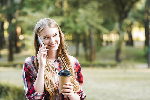selective focus of beautiful girl in casual clothes holding paper coffee cup while talking on smartphone and looking away