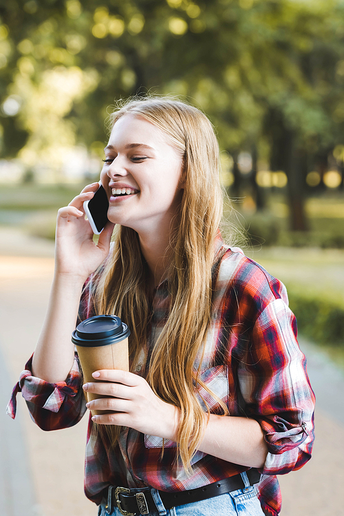 portrait shot of beautiful girl in casual clothes holding paper coffee cup and laughing with closed eyes while talking on smartphone