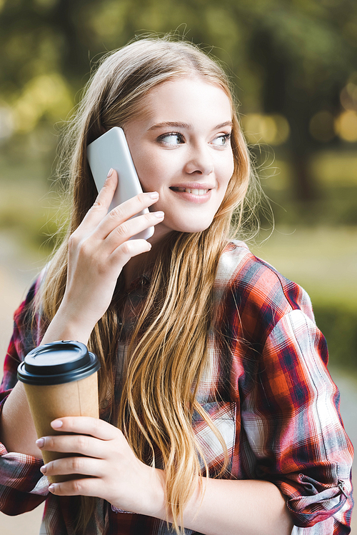 portrait shot of beautiful girl in casual clothes holding paper coffee cup and smiling while talking on smartphone and looking away