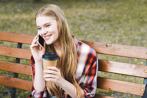 beautiful girl in casual clothes holding paper coffee cup while sitting on wooden bench in park and talking on smartphone